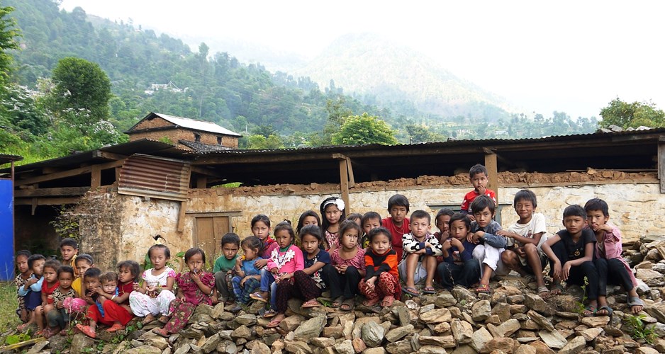 A group of children of all ages sit on a pile of rubble in front of a building, in a rural part of Nepal.
